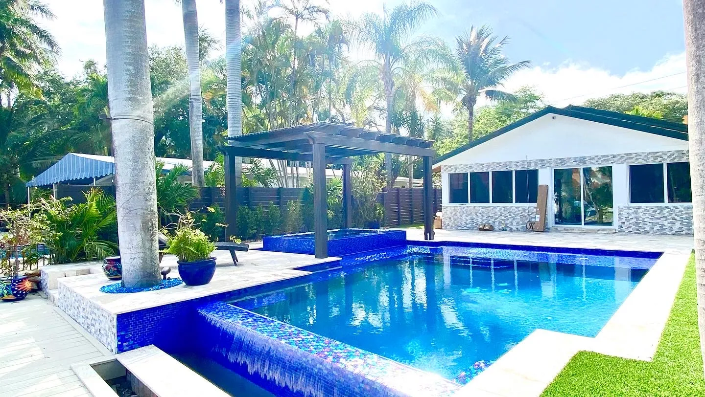 Pool with iridescent blue glass tile and spillover edge, raised spa under a bronze pergola and artificial turf