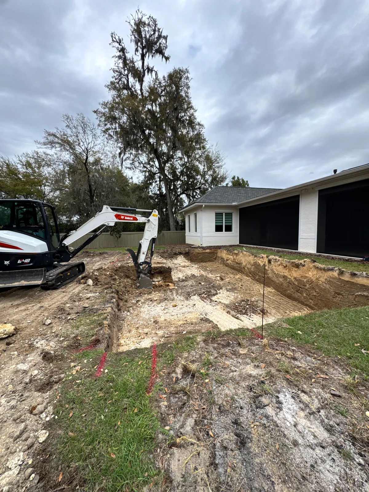 Mini excavator digging a new pool beside a white brick home with a screened patio.