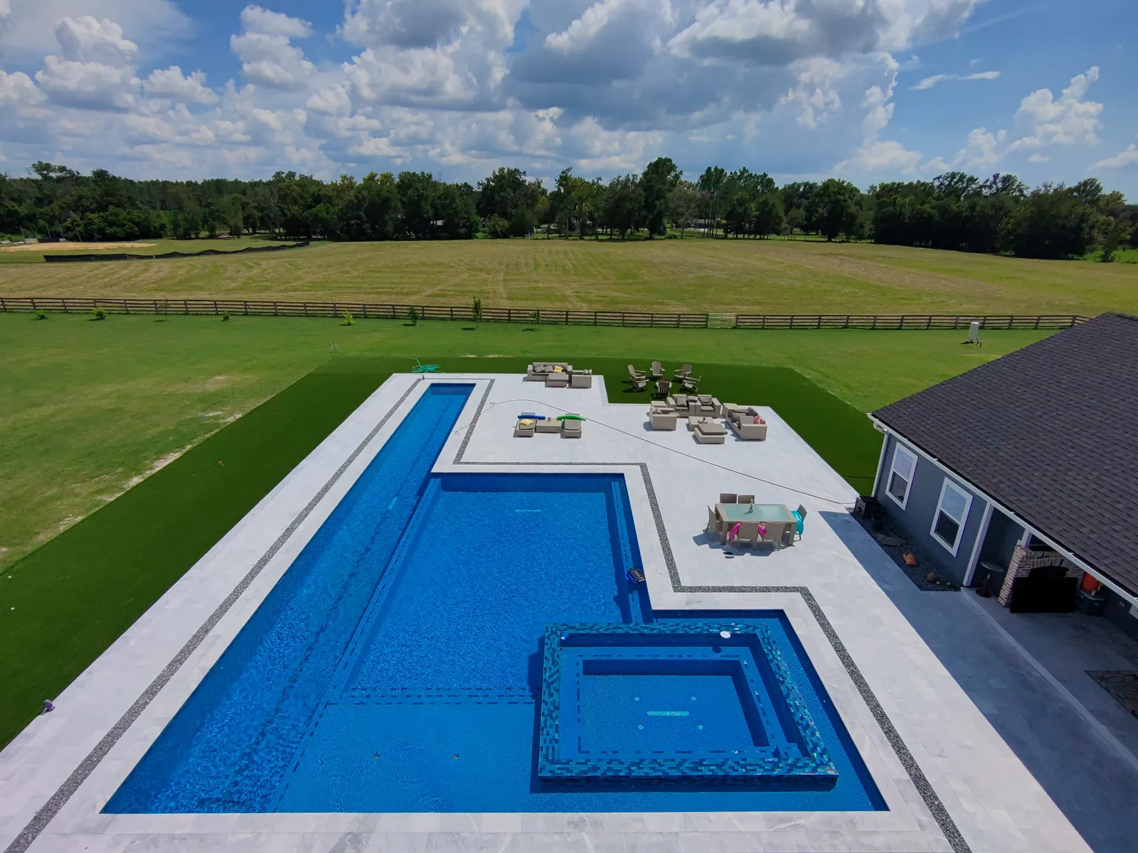 Aerial view of an L-shaped pool with square raised spa, gray marble deck and turf border on a rural property.