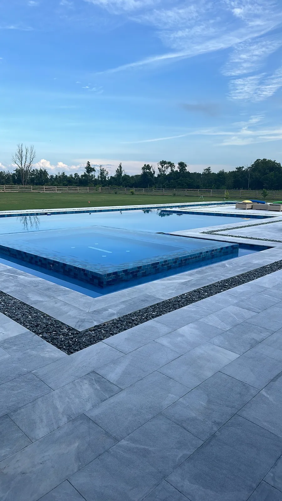 Raised tiled spa inside a new pool with gray marble deck and pebble band, open pasture behind at dusk.