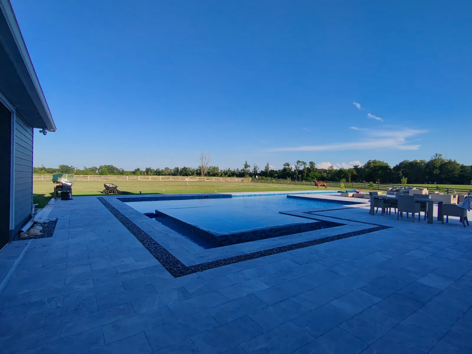 Raised spa and pool edged with a black pebble band in a wide gray marble deck under a clear evening sky.