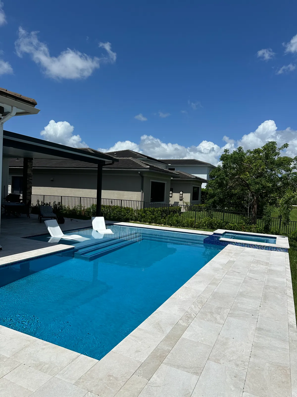 Rectangular pool with raised spa, sun shelf loungers and ivory travertine deck under a blue sky