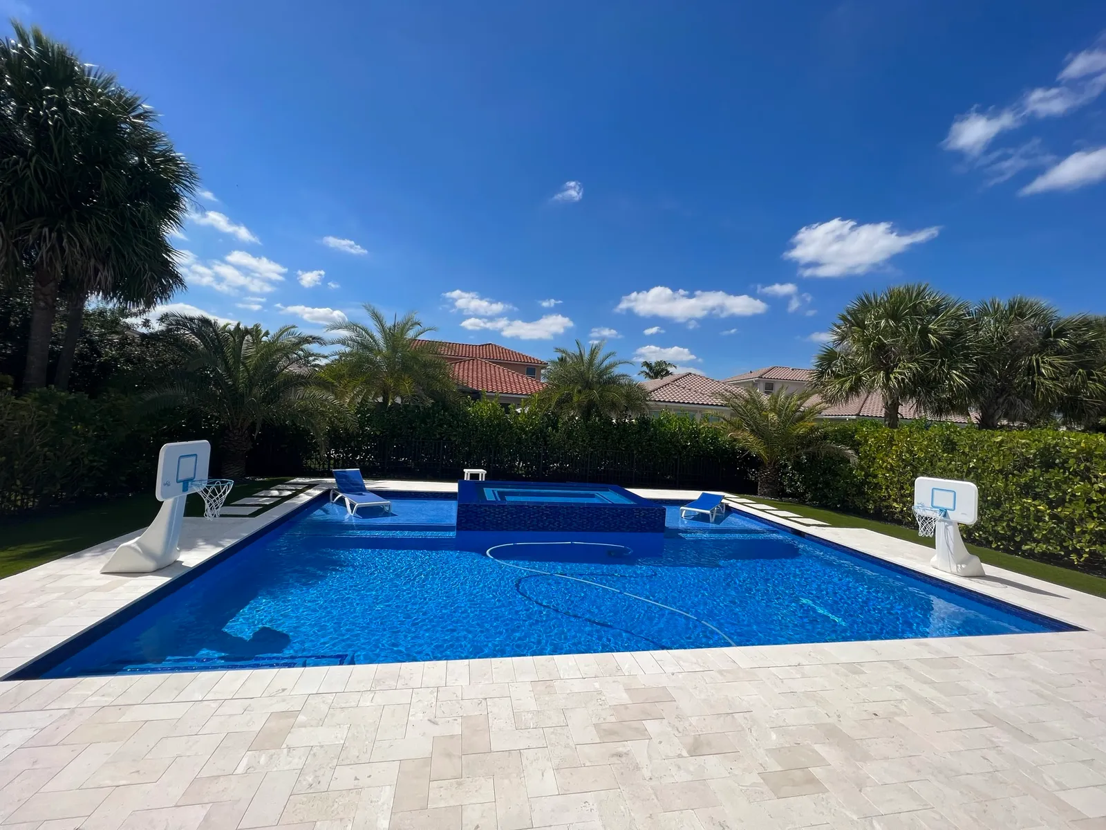 Wide view of a new pool with raised spa and ivory travertine deck framed by hedges and palm trees.