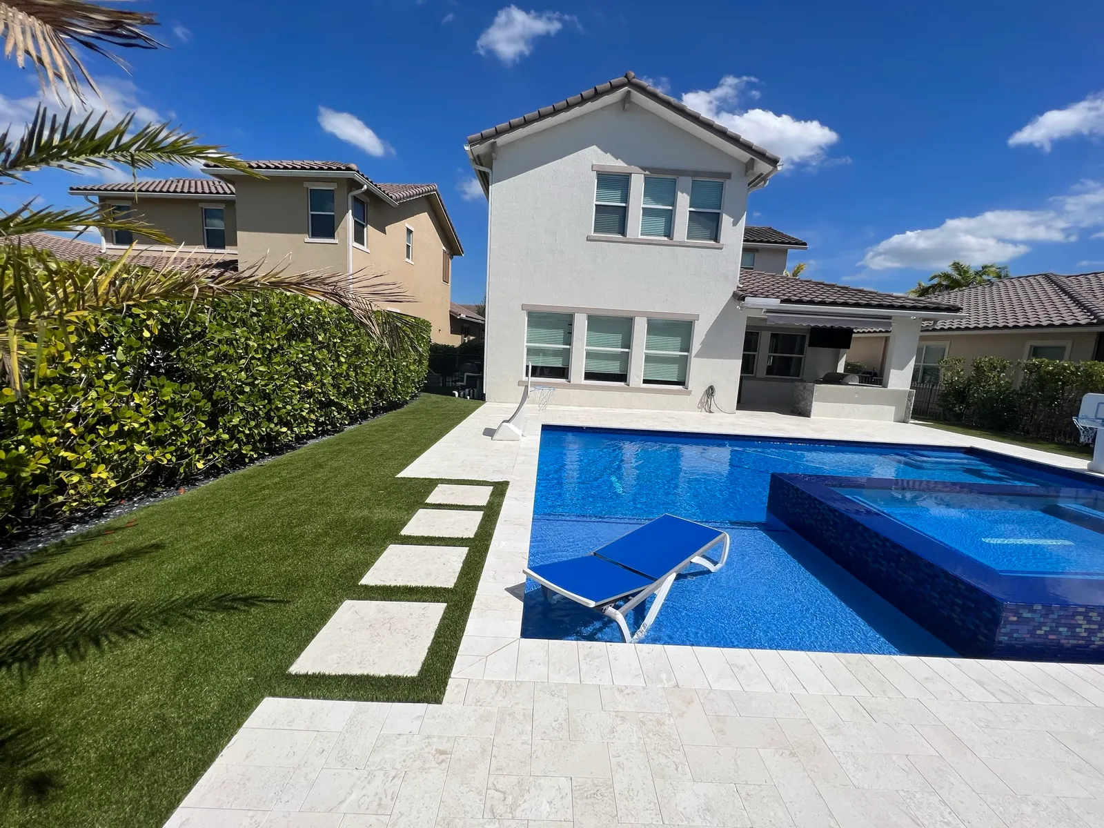 New pool with raised spa, travertine deck and stepping pads in artificial turf behind a two-story home.