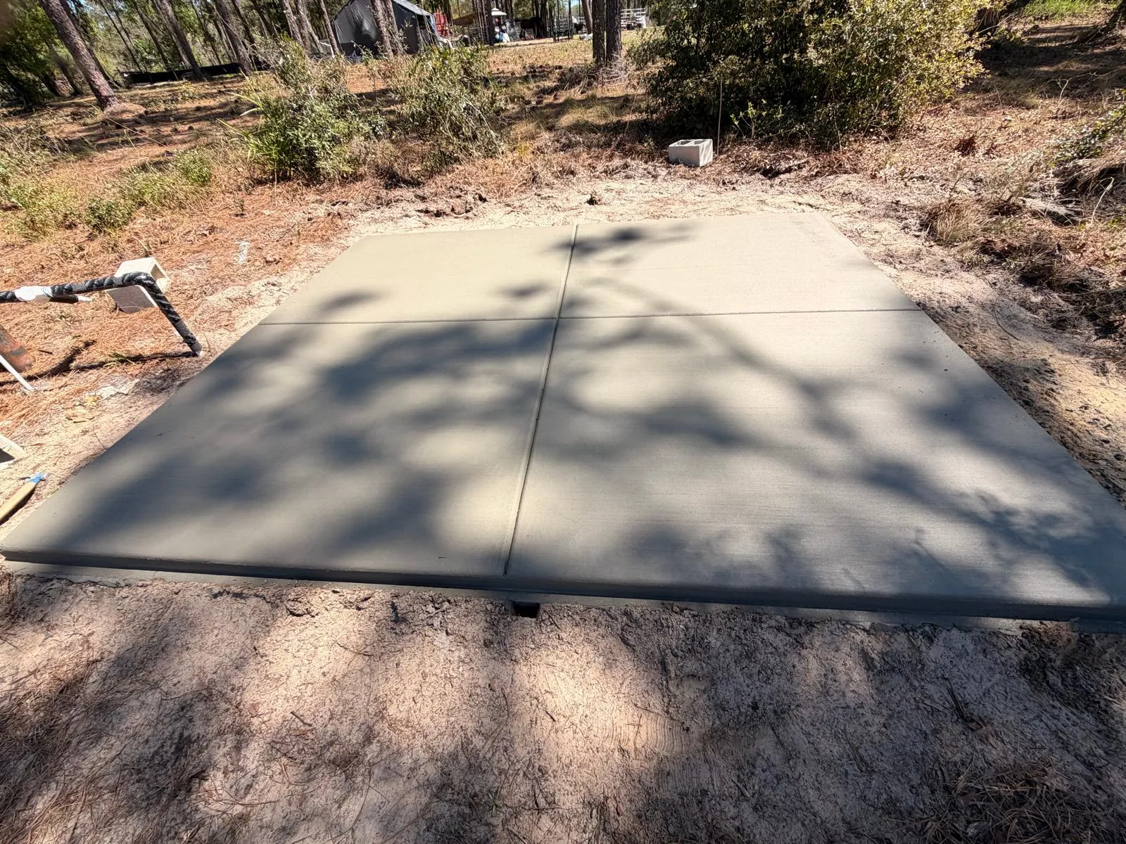 Square concrete pad with cross control joints poured in a wooded lot.