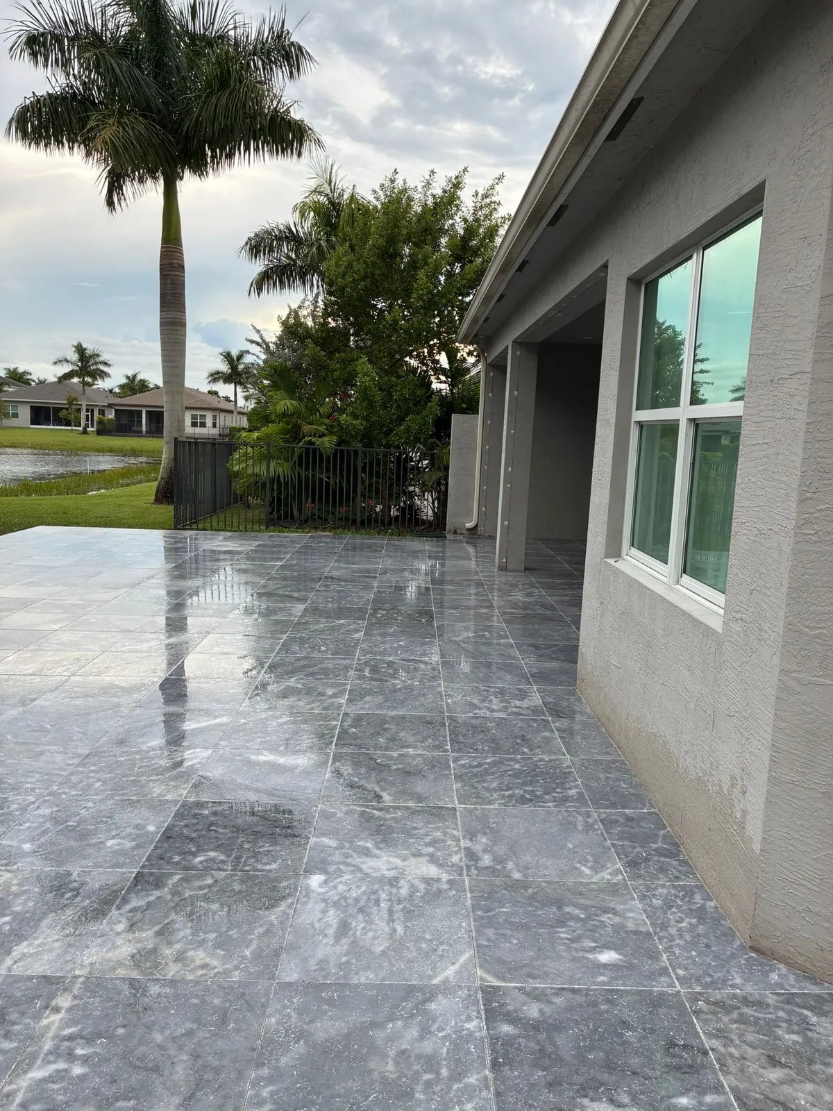 Wet gray marble paver patio along a stucco house with a royal palm and lake behind.