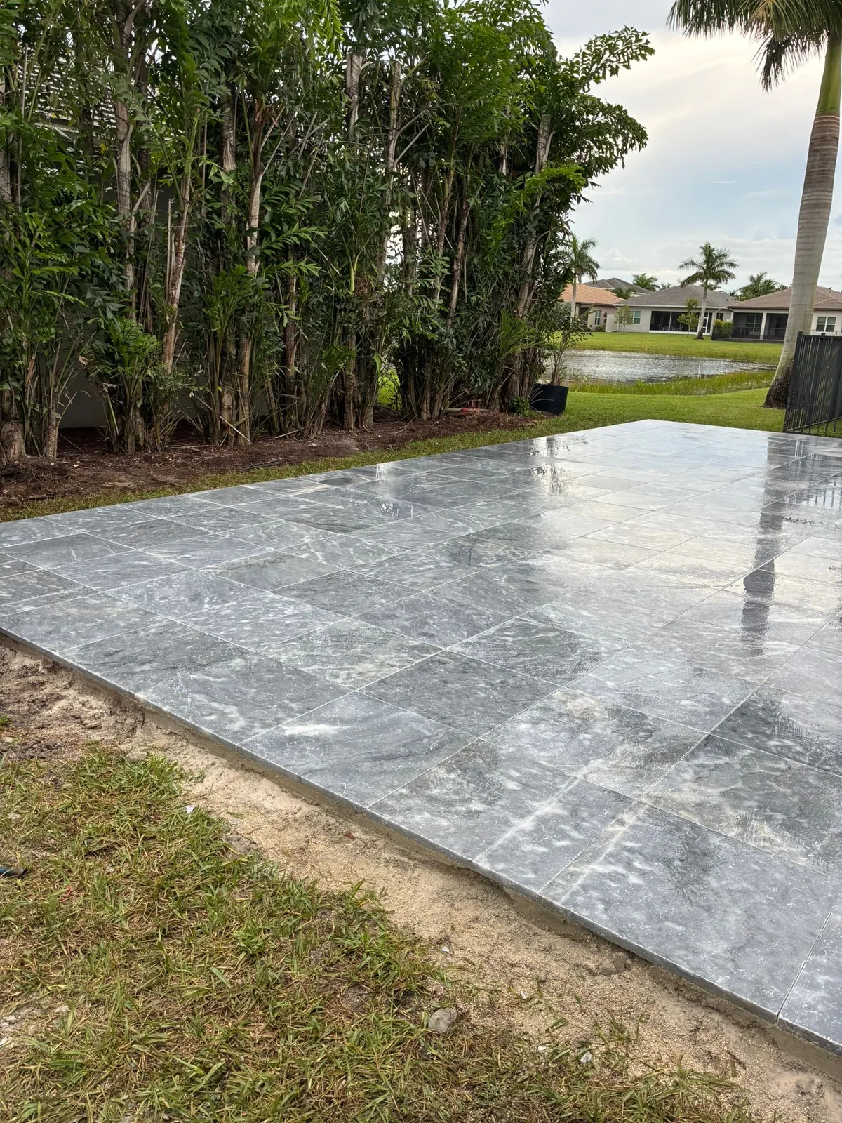 Edge of a new gray marble paver patio beside a bamboo palm hedge and lake.