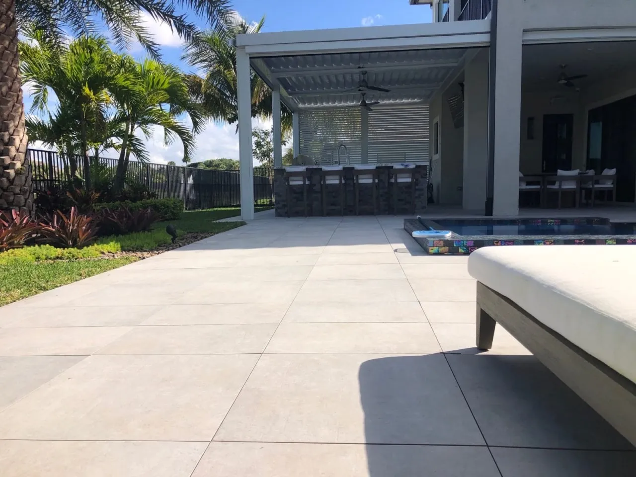 Large-format porcelain pool deck leading to a white louvered pergola and outdoor kitchen.