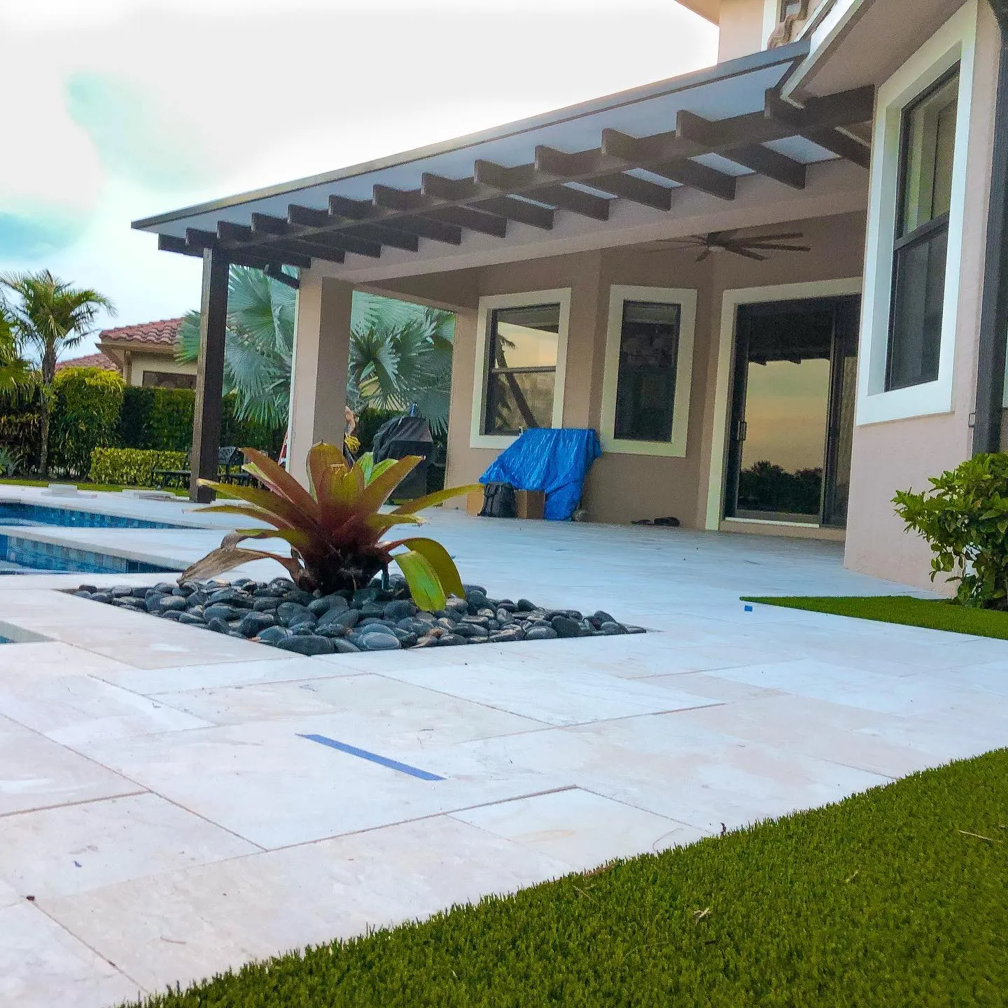 Ivory travertine pool deck with a bromeliad in black river rock, artificial turf and a pergola-style patio cover