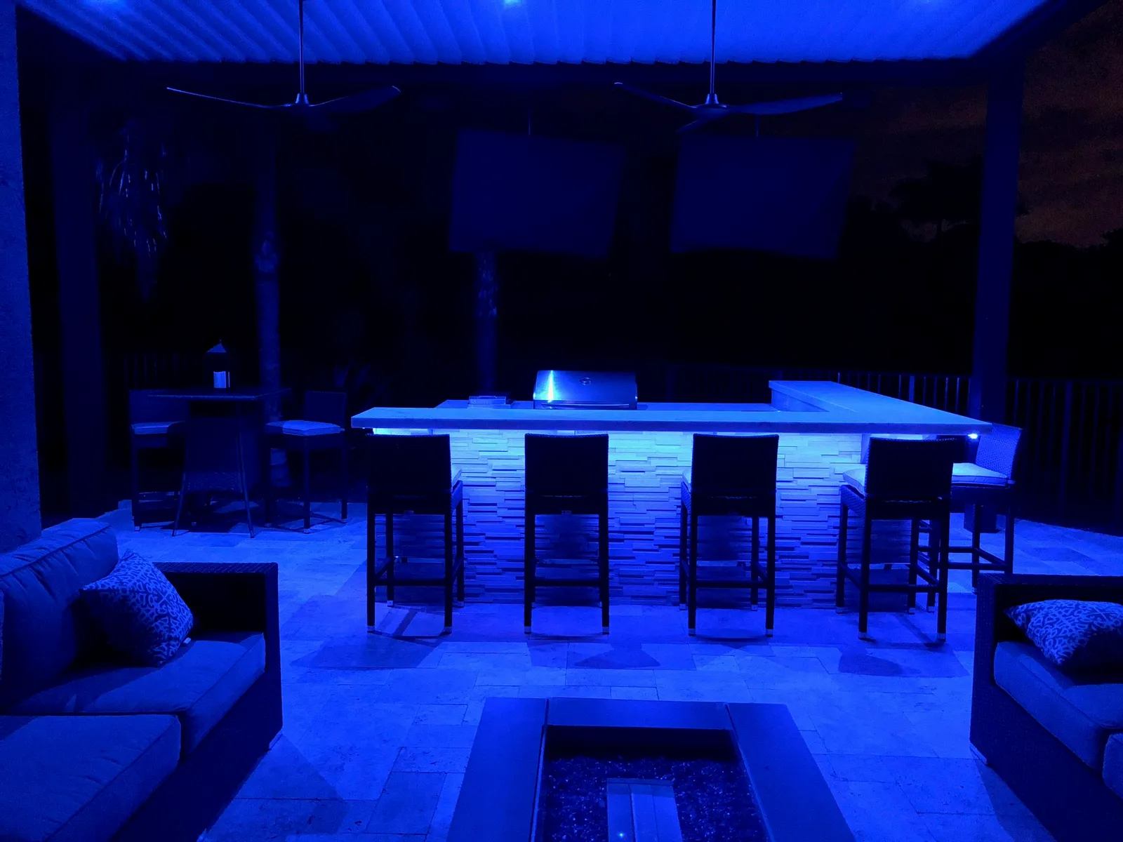 Outdoor kitchen bar lit with blue LED lighting at night under a louvered roof with a fire pit.