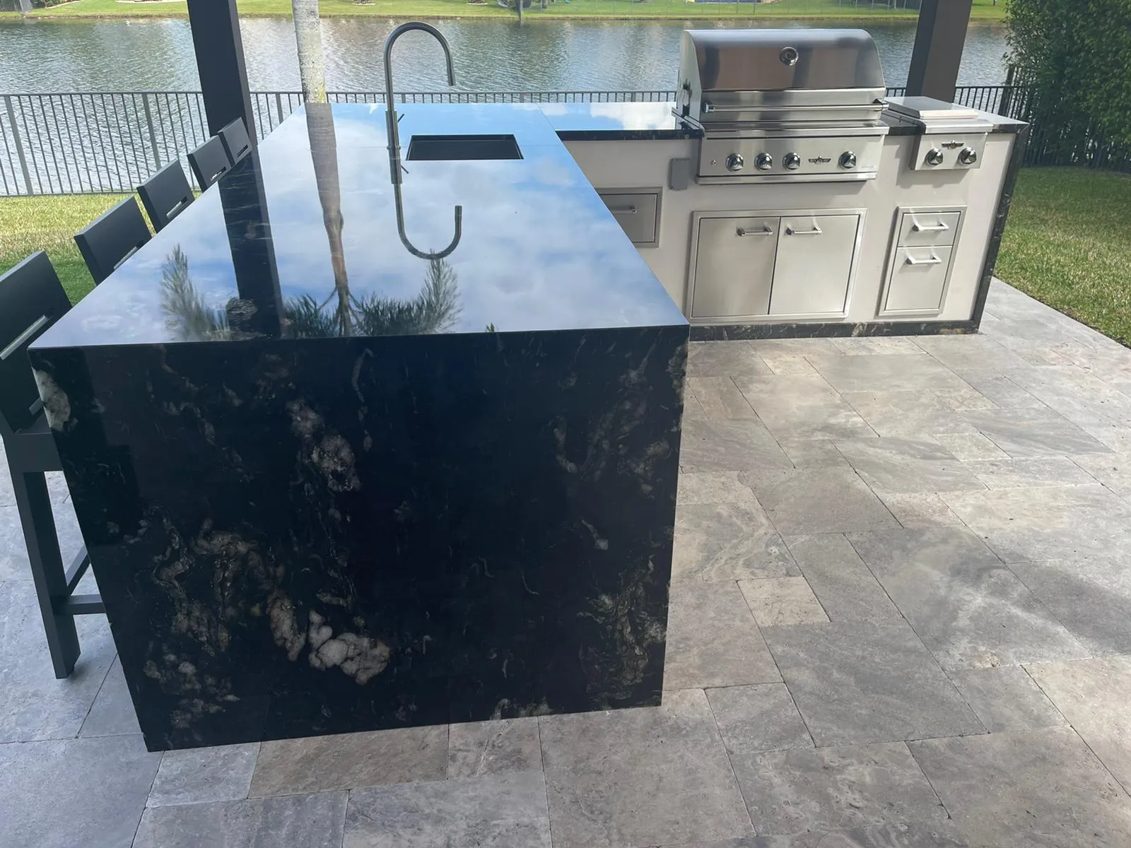 Black granite waterfall island with sink in front of a built-in grill station by a lake.