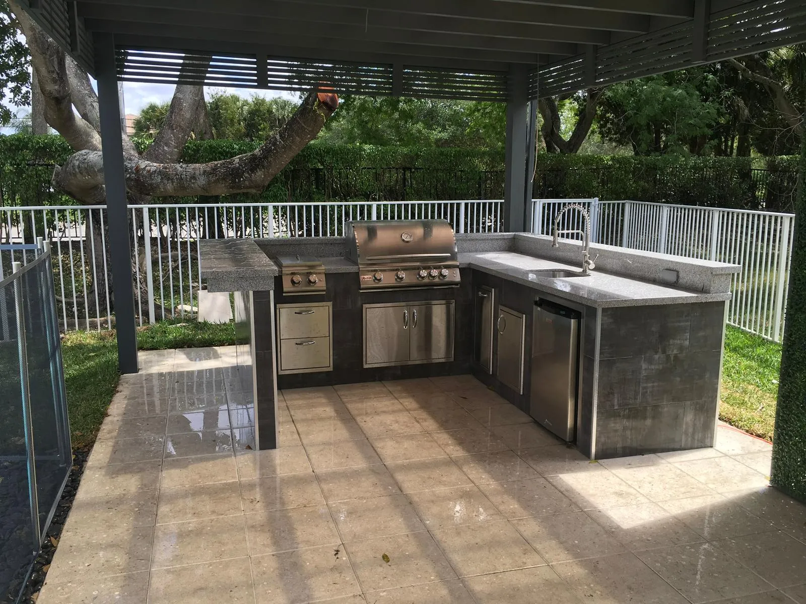 U-shaped outdoor kitchen with gray tile, granite countertop and grill under a pergola.