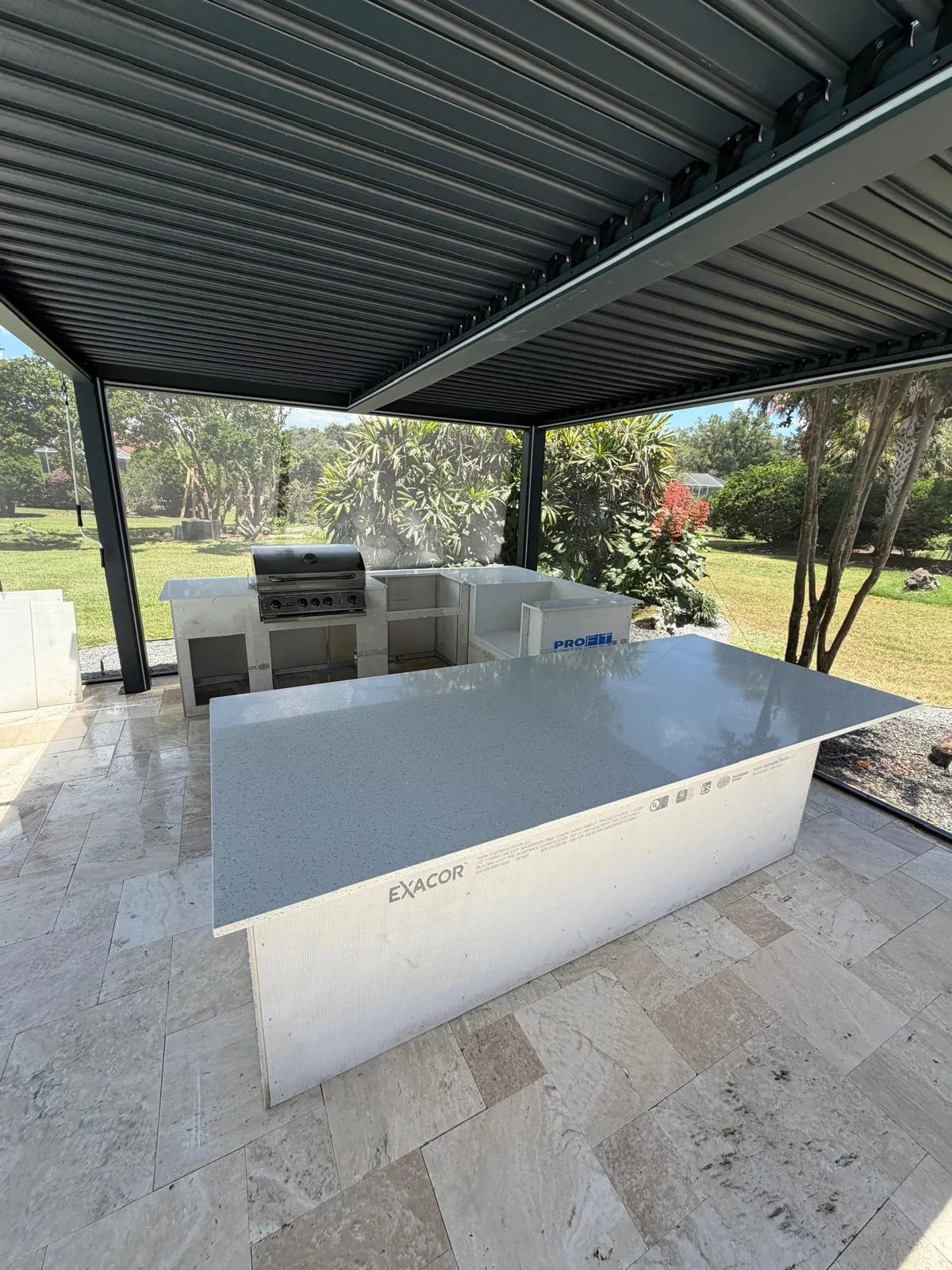 Large quartz-topped bar island and grill station under a louvered pergola in a backyard.