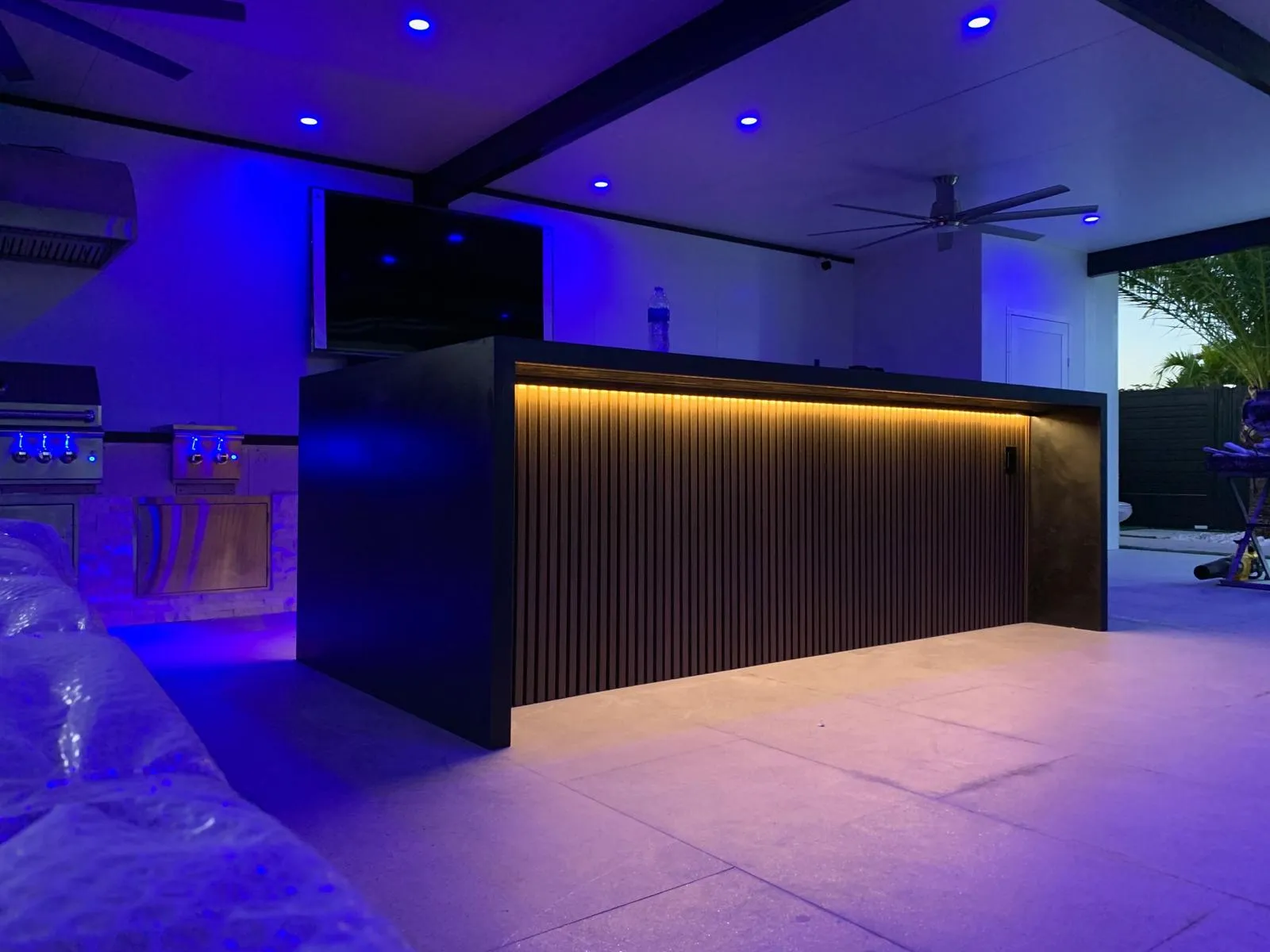 Outdoor kitchen island with slatted front and warm LED strip under blue ceiling lights at dusk.