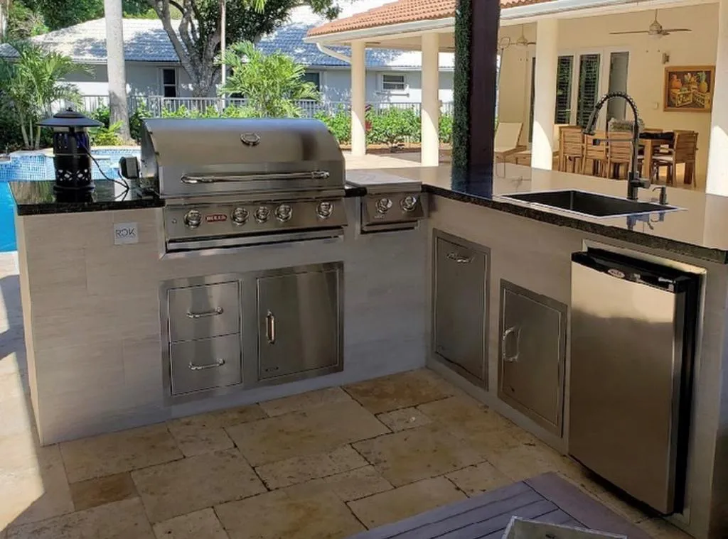 L-shaped tiled outdoor kitchen with black granite top, grill, sink and fridge beside a pool.