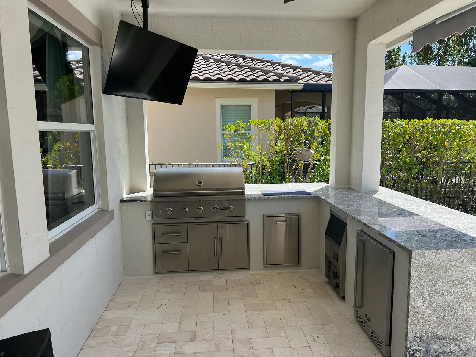 L-shaped outdoor kitchen with built-in grill, ice maker, fridge and granite countertop on a covered patio.