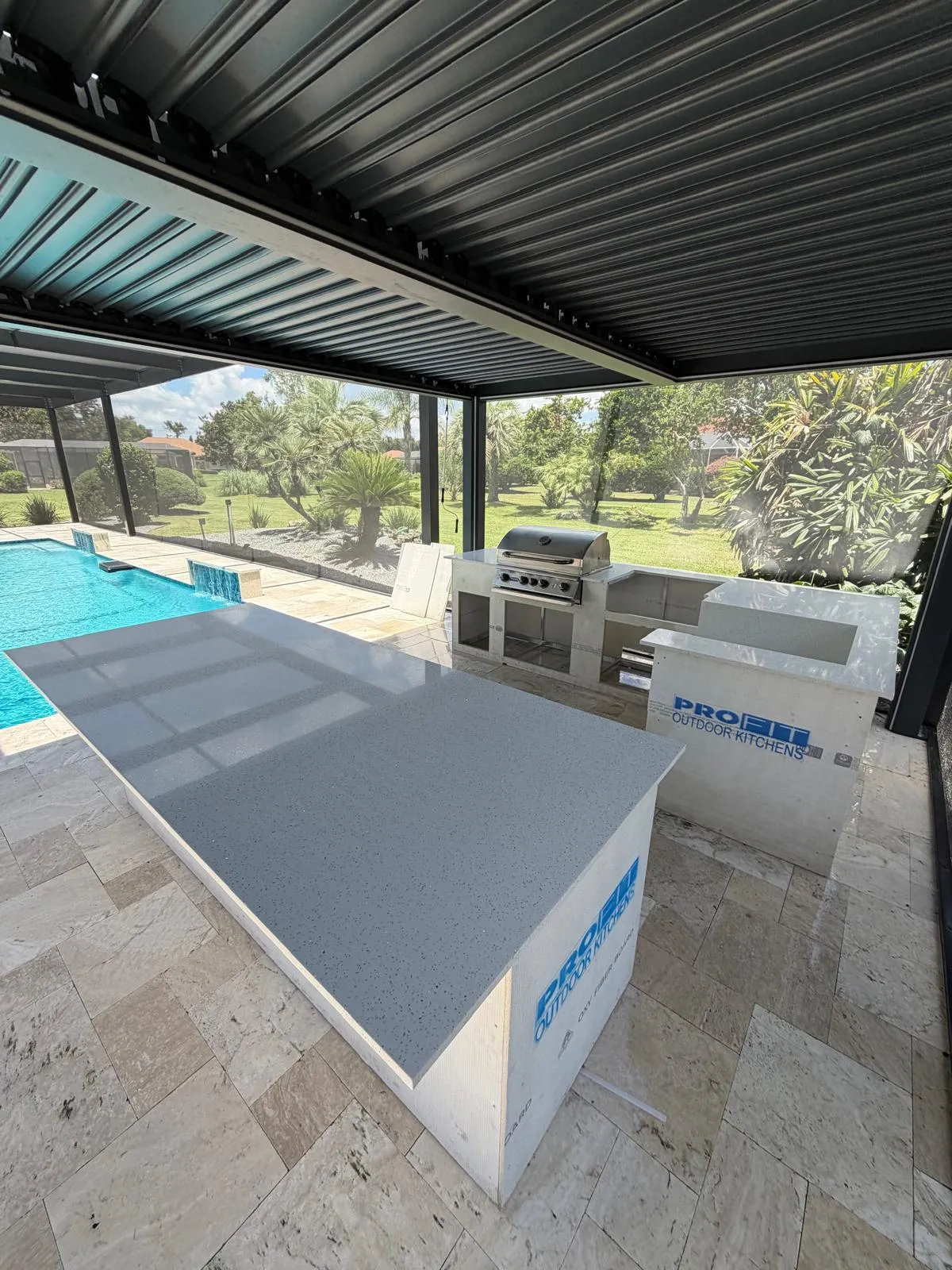 Poolside outdoor kitchen with gray quartz bar under a louvered pergola, pool waterfalls behind.