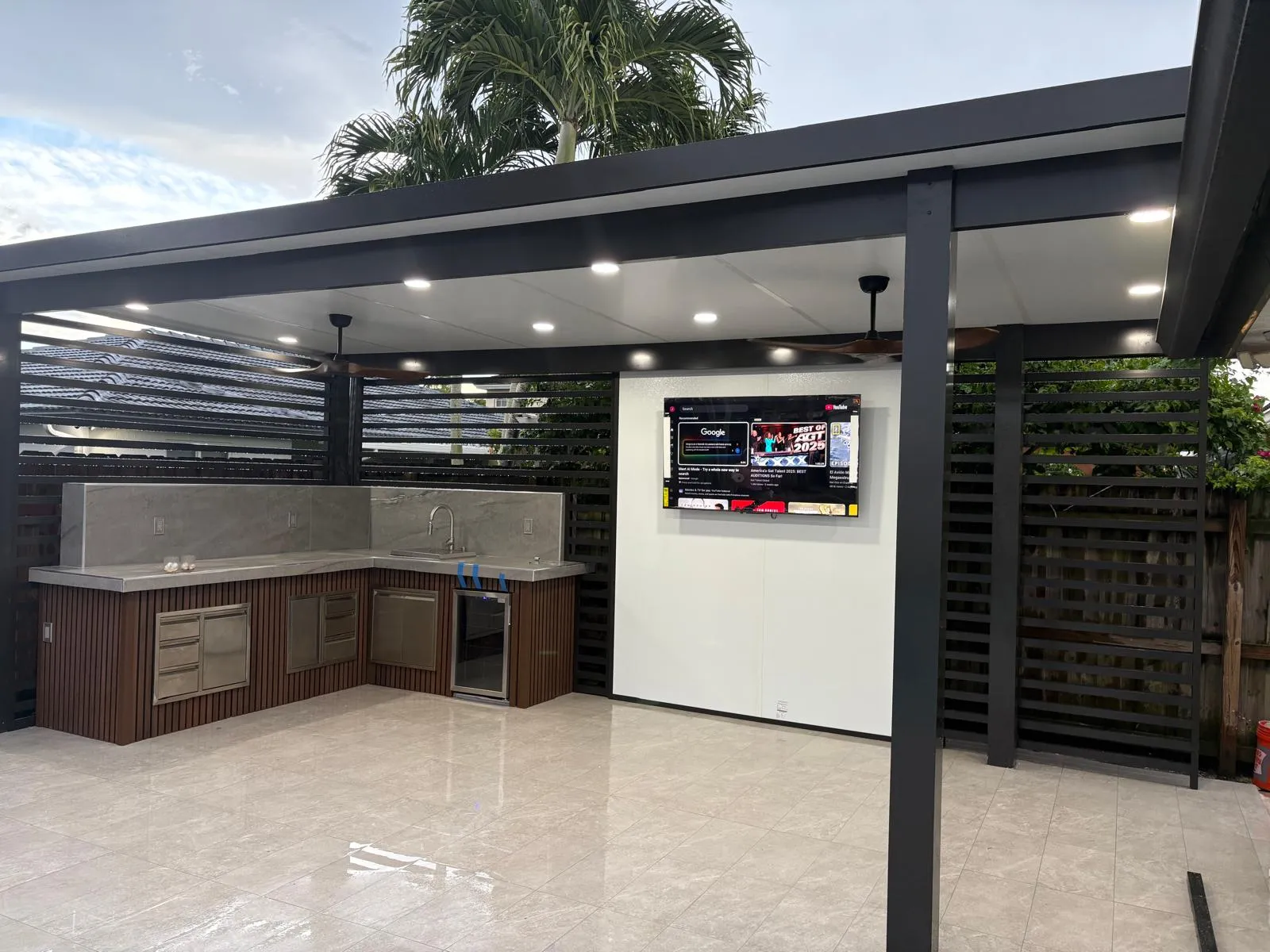 L-shaped outdoor kitchen with slatted wood base, sink and wine fridge under a black-framed patio cover with TV