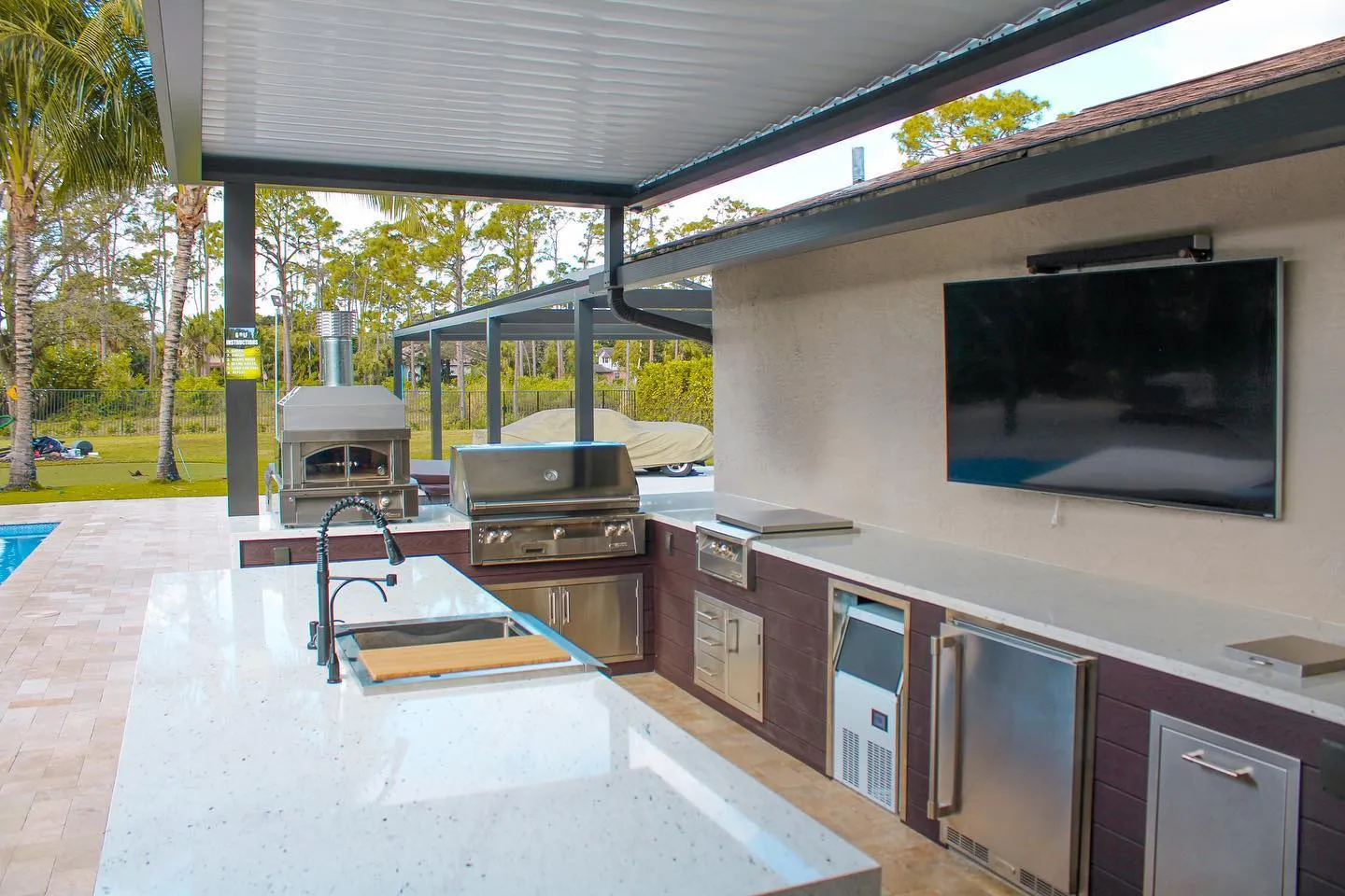 Outdoor kitchen with pizza oven, built-in grill, sink and wall TV under a louvered roof.
