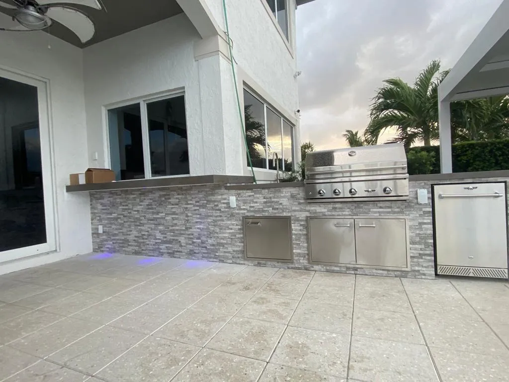 Gray stacked stone outdoor kitchen with built-in grill and refrigerator at dusk.