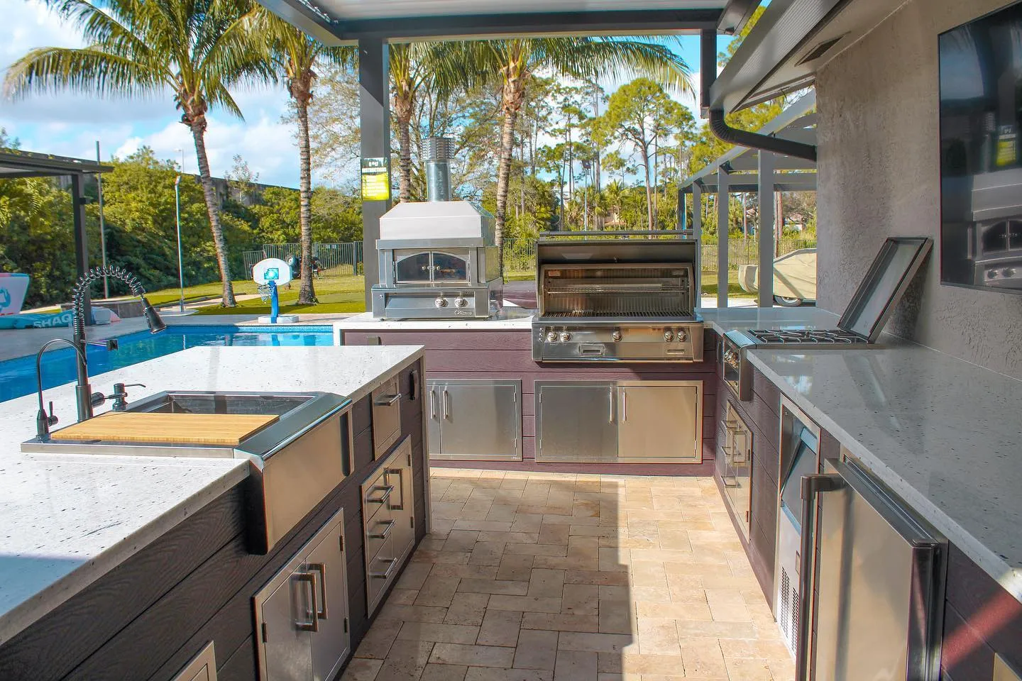 U-shaped outdoor kitchen with pizza oven, open grill and side burner beside a pool.