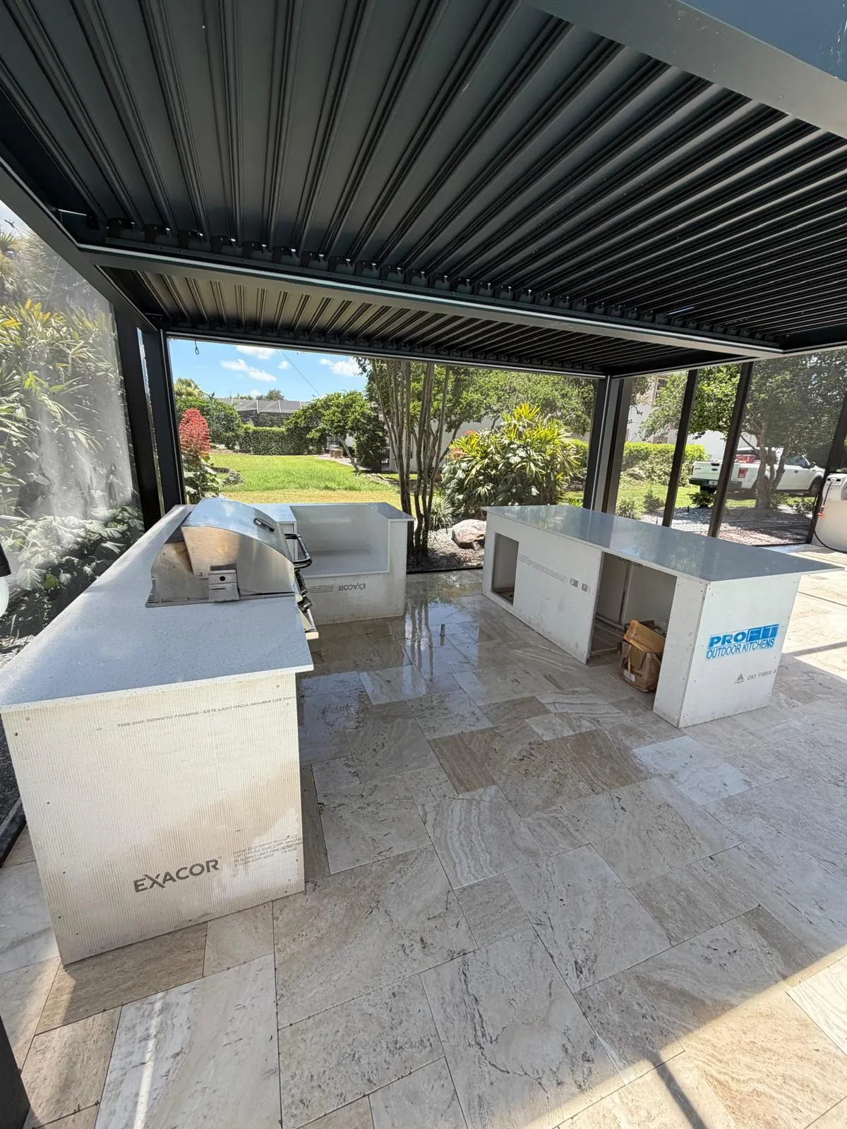 Outdoor kitchen islands with quartz tops and grill being built under a dark louvered pergola.