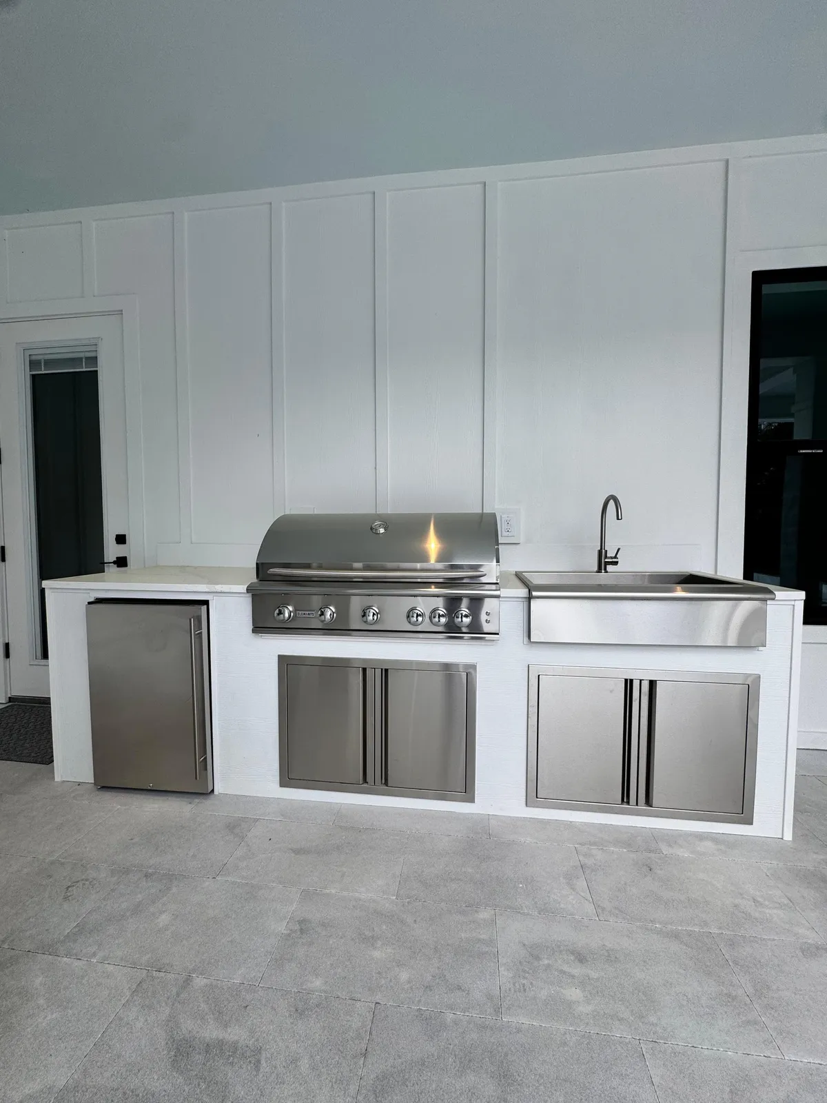 White straight-run outdoor kitchen with built-in grill, farmhouse sink and fridge on a lanai.