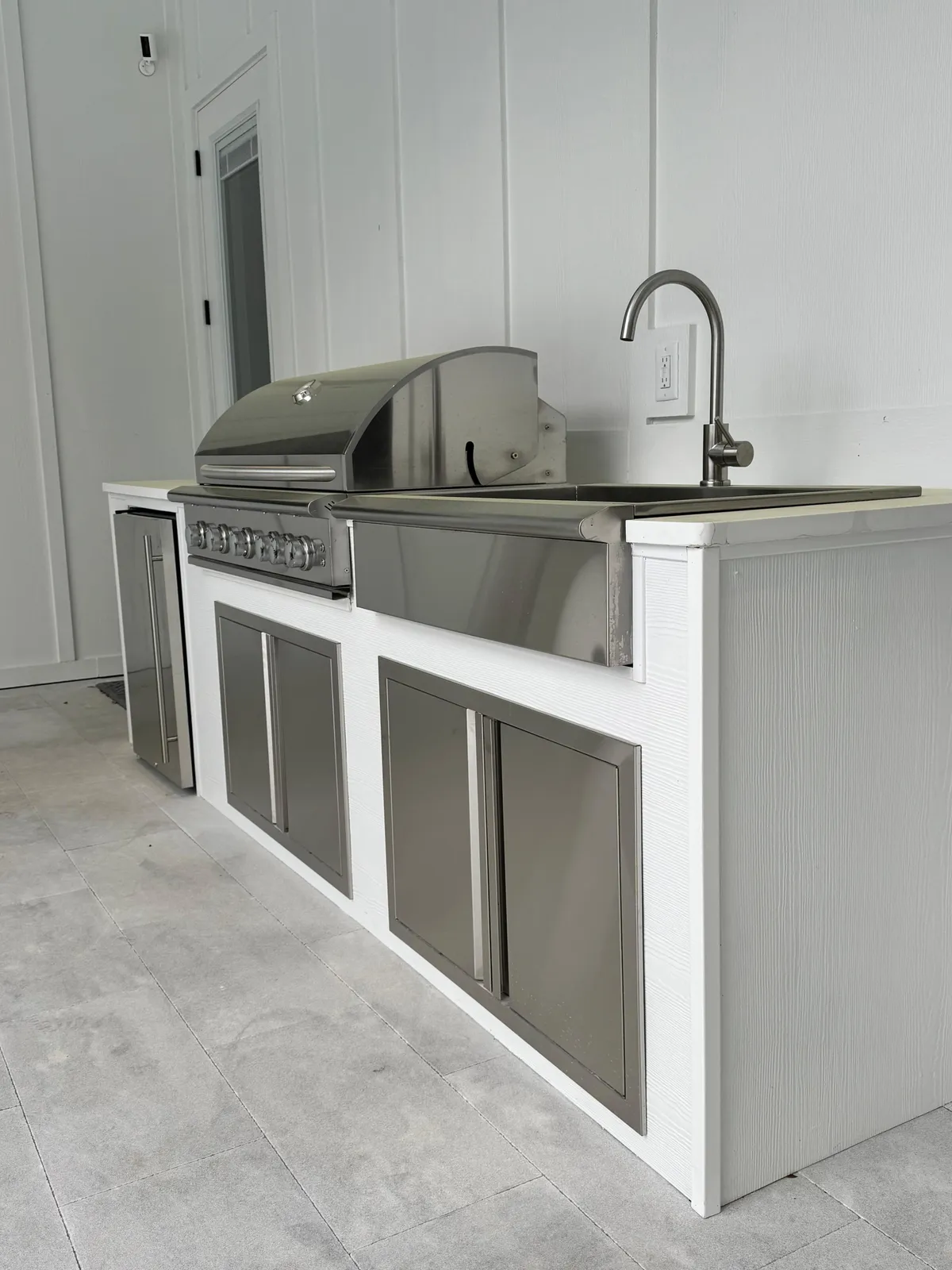 Side view of a white outdoor kitchen with stainless farmhouse sink, grill and double access doors.