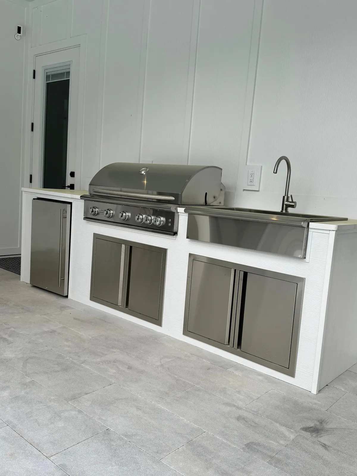White outdoor kitchen with built-in grill, farmhouse sink and under-counter fridge on gray pavers.
