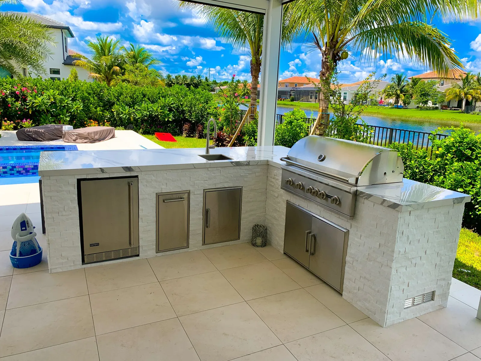 L-shaped outdoor kitchen in white stacked stone with built-in grill, sink and lake view.