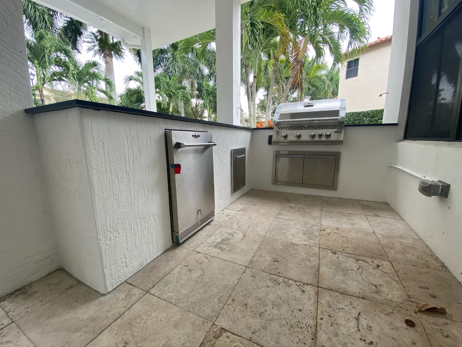 White stucco outdoor kitchen interior with built-in grill, refrigerator and travertine floor.