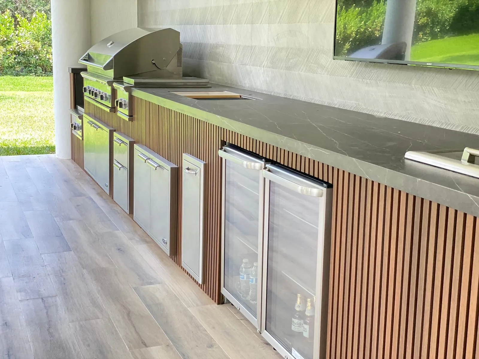 Wood slat outdoor kitchen with glass-door beverage fridges and dark stone countertop.