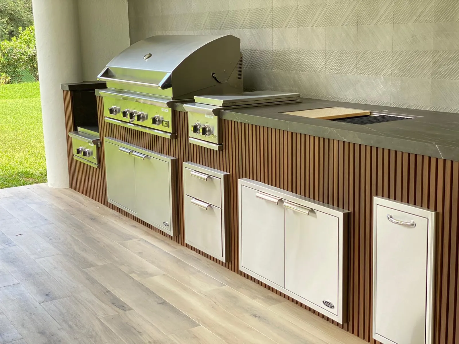 Outdoor kitchen with vertical wood slat cladding, stainless grill, side burner and access doors.
