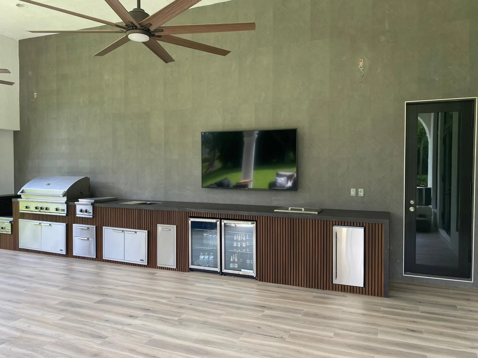 Long wood slat outdoor kitchen with grill, beverage fridges and wall TV under a large ceiling fan.