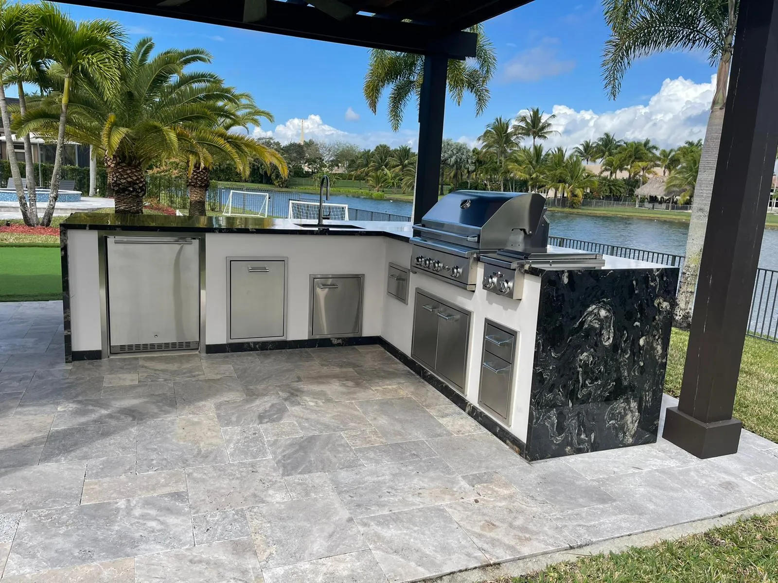 L-shaped outdoor kitchen with black granite waterfall under a dark pergola beside a lake.