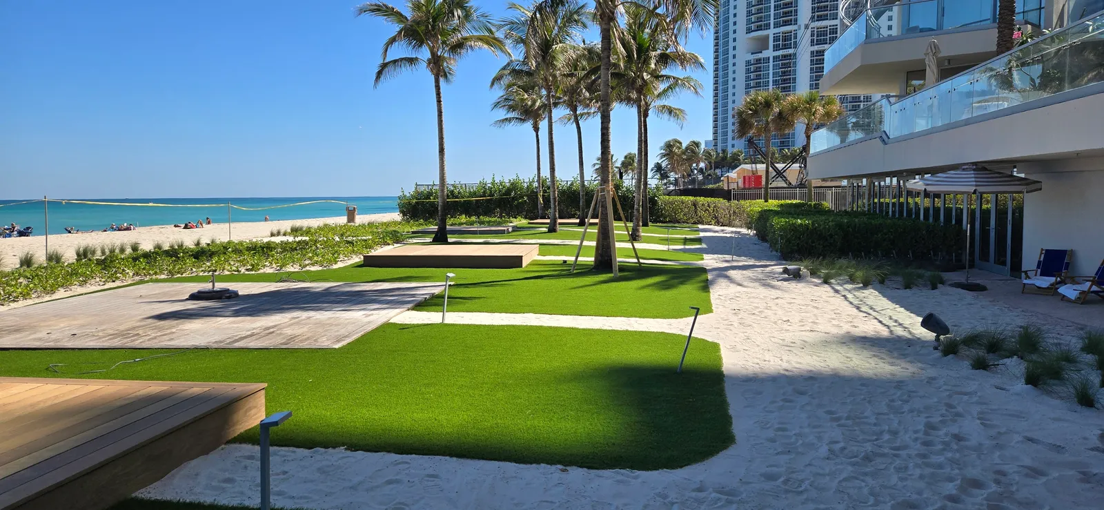 Artificial turf lawns and wood deck platforms between sand paths at a beachfront resort.