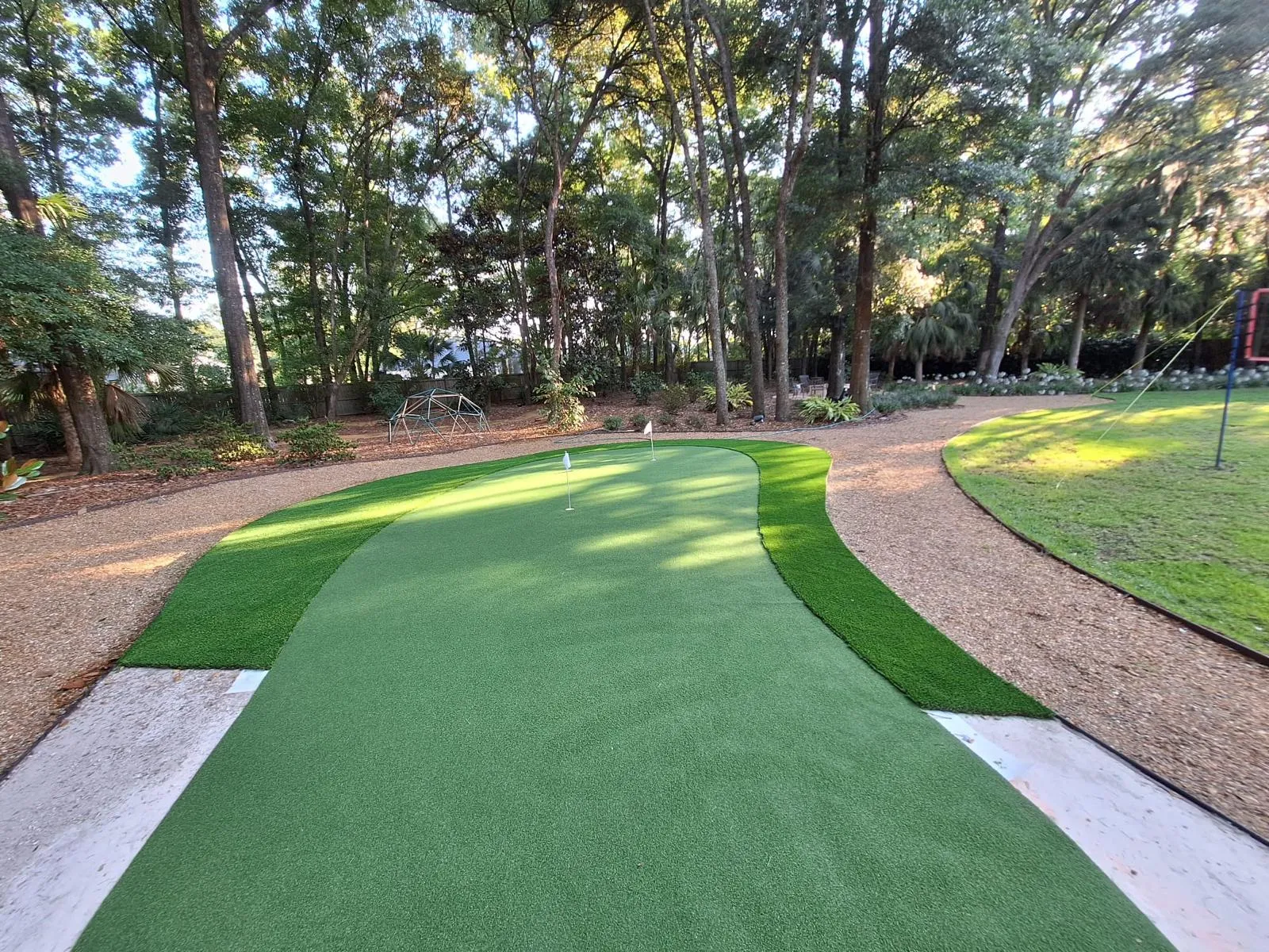 Kidney-shaped artificial turf putting green with fringe under tall oak trees and gravel paths.