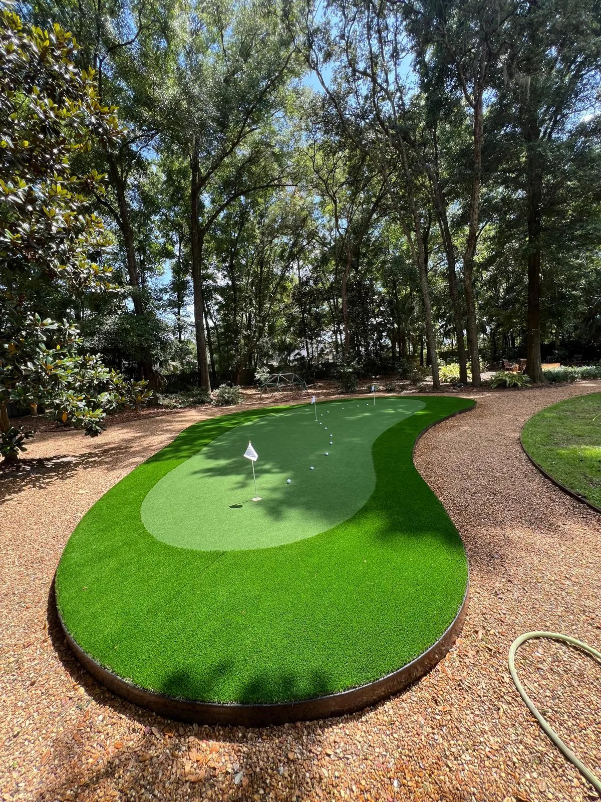 Finished kidney-shaped putting green with fringe turf and steel edging under oak trees.