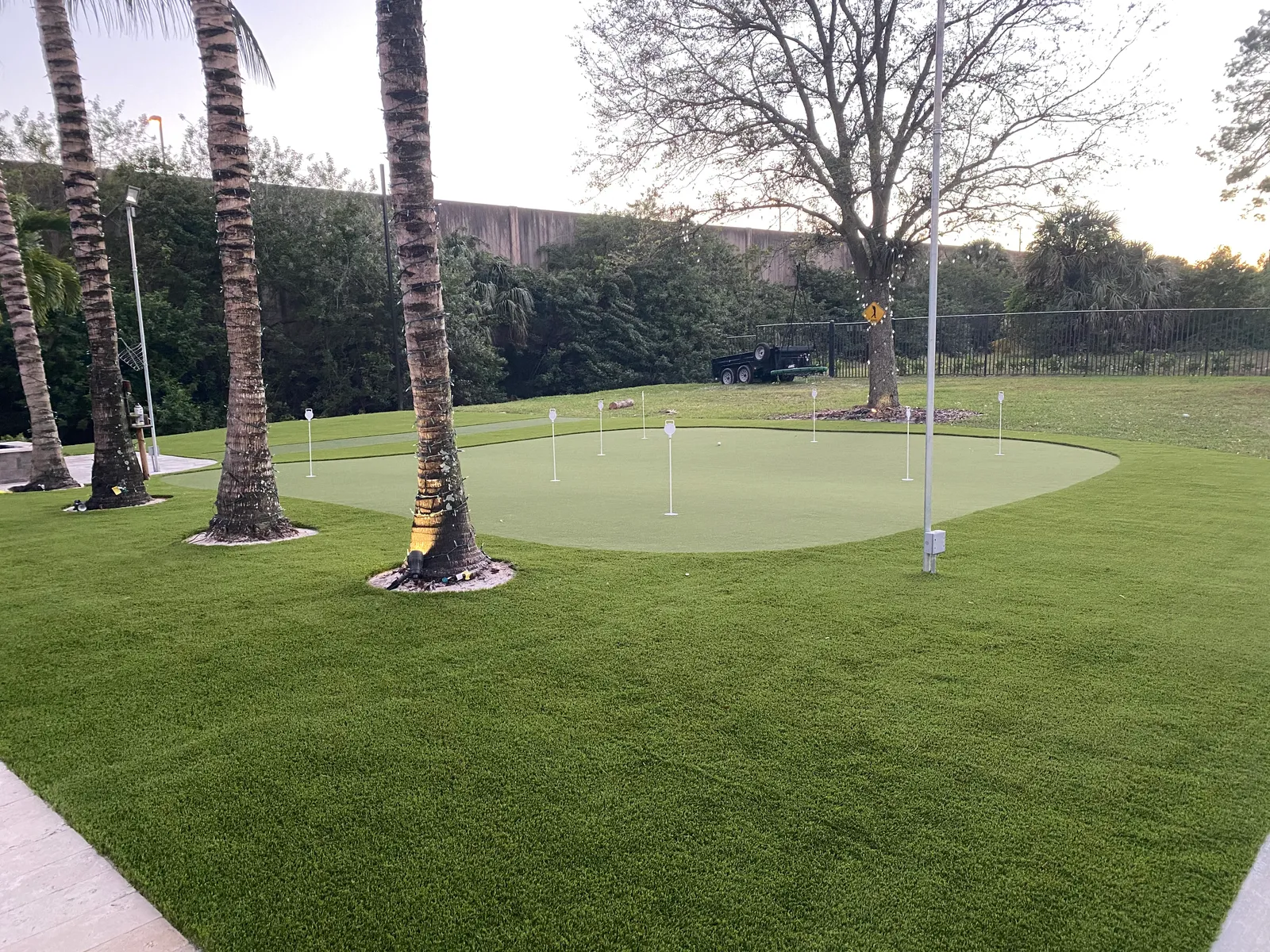 Backyard putting green at dusk with uplights on palm trunks and wide artificial turf lawn.