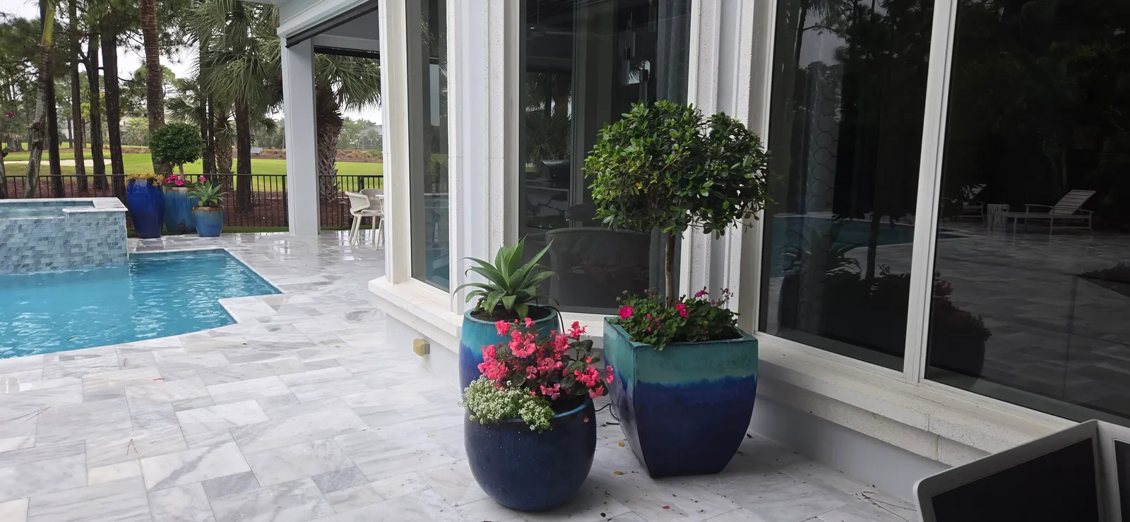 Blue glazed planters with a topiary, agave and pink flowers on a marble pool deck.