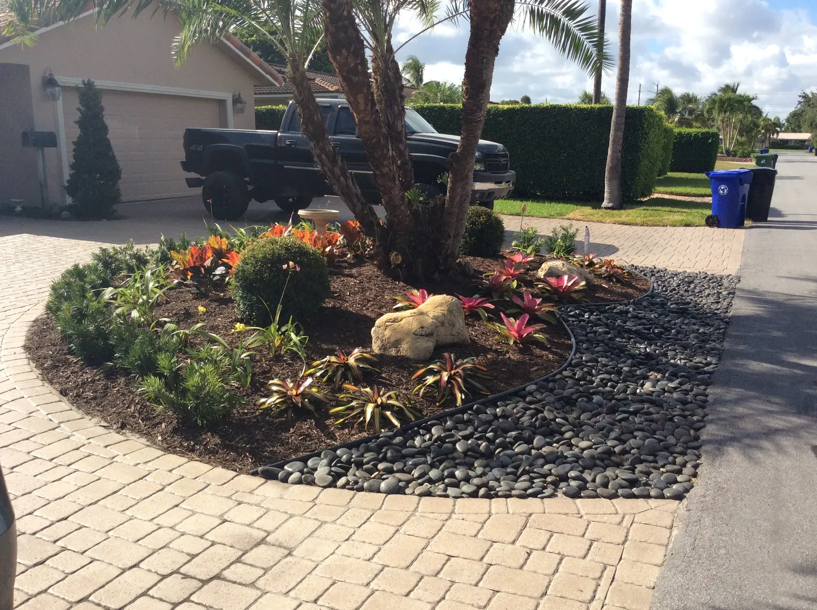 Driveway island with bromeliads, crotons and boulders edged by black river rock and pavers.