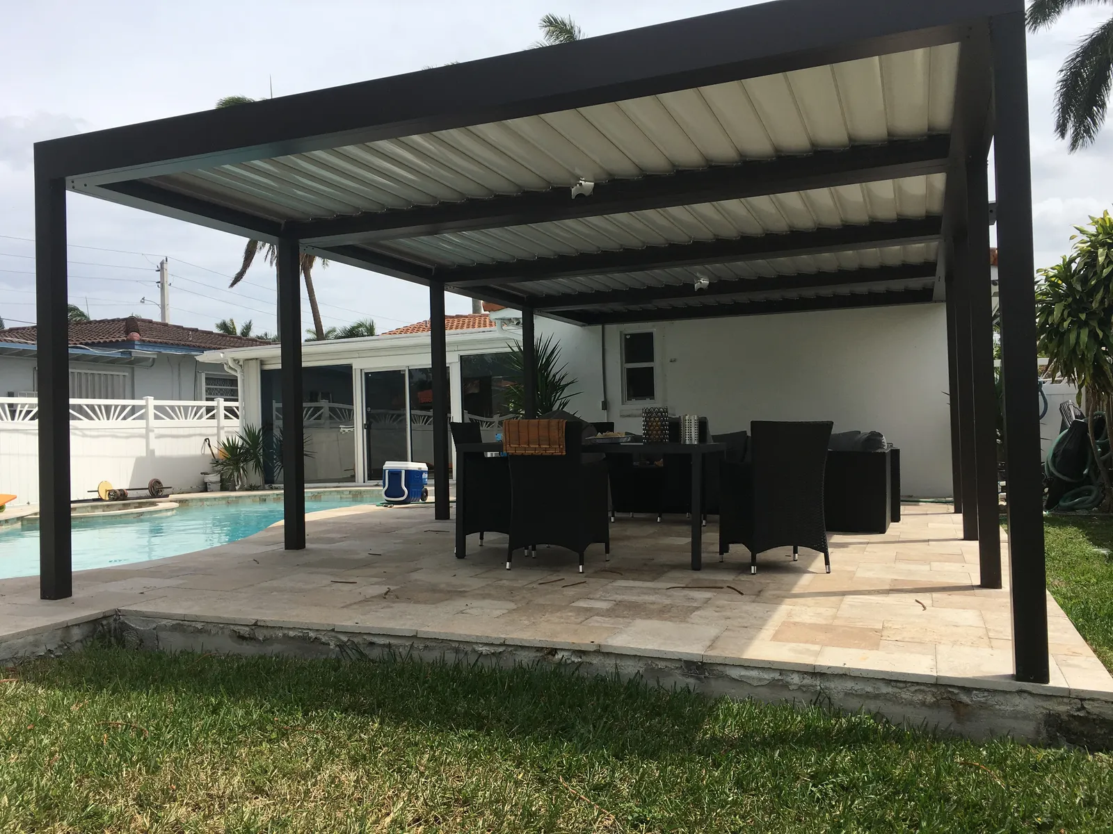 Bronze louvered pergola with closed roof over a wicker dining set on a travertine pool deck