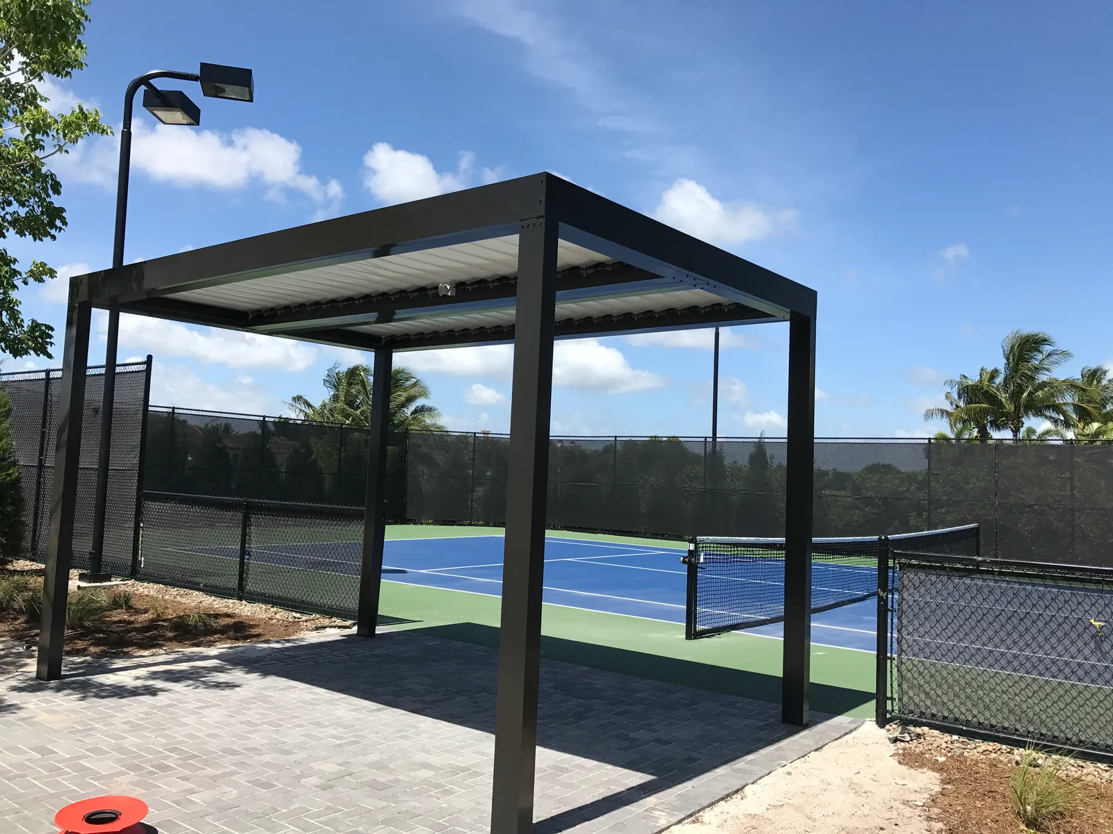 Bronze freestanding louvered pergola on a paver pad beside a blue tennis court
