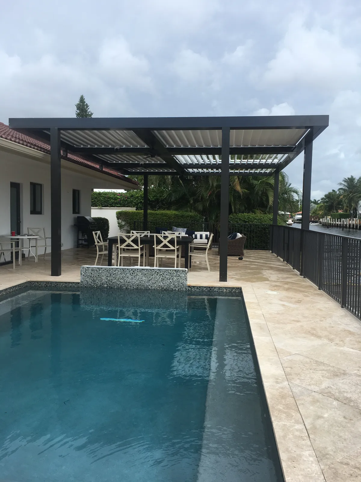 Bronze louvered pergola over a dining area on a travertine pool deck beside a canal