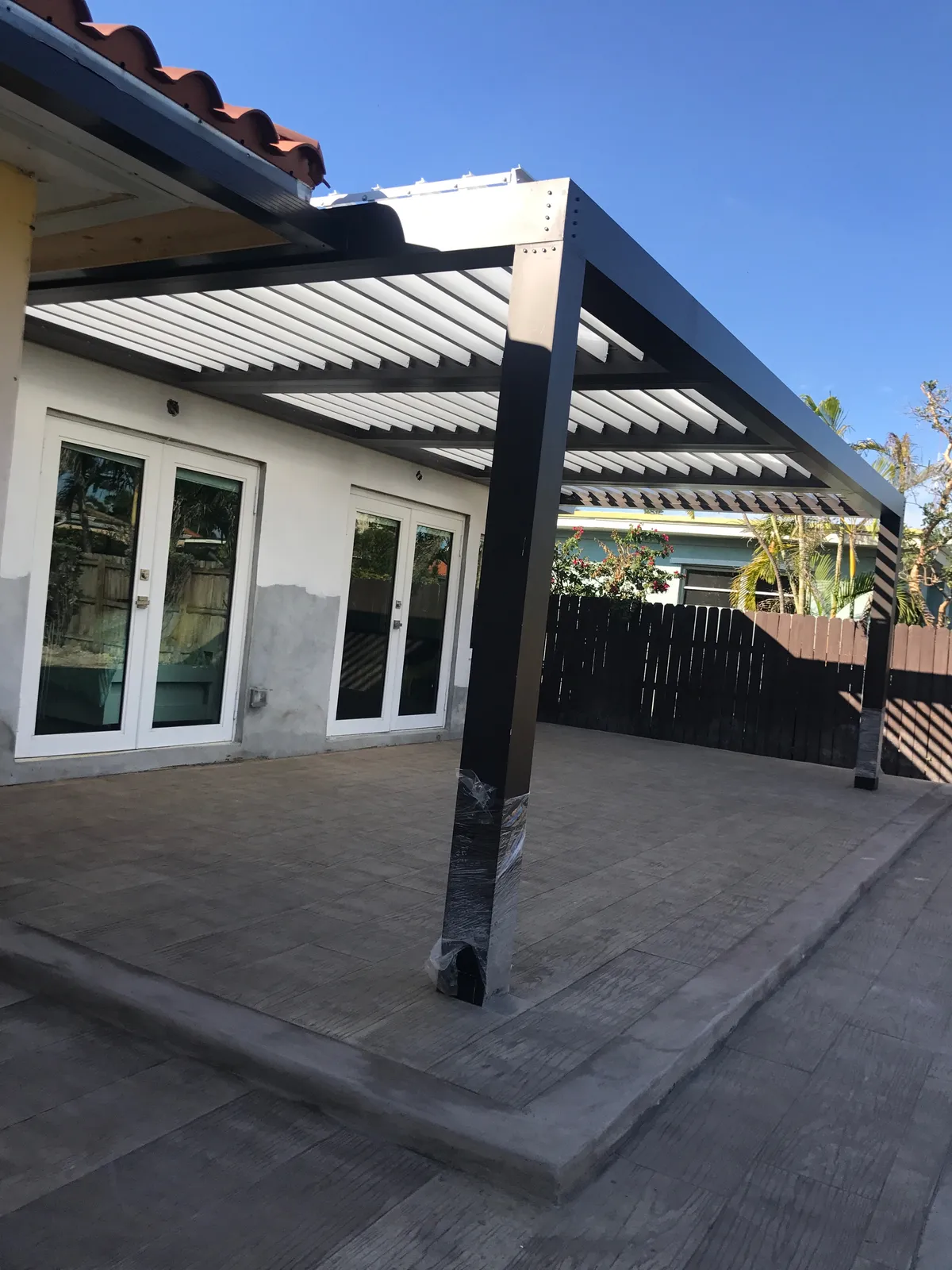 Newly installed bronze louvered pergola with posts still wrapped in protective film over a stamped patio