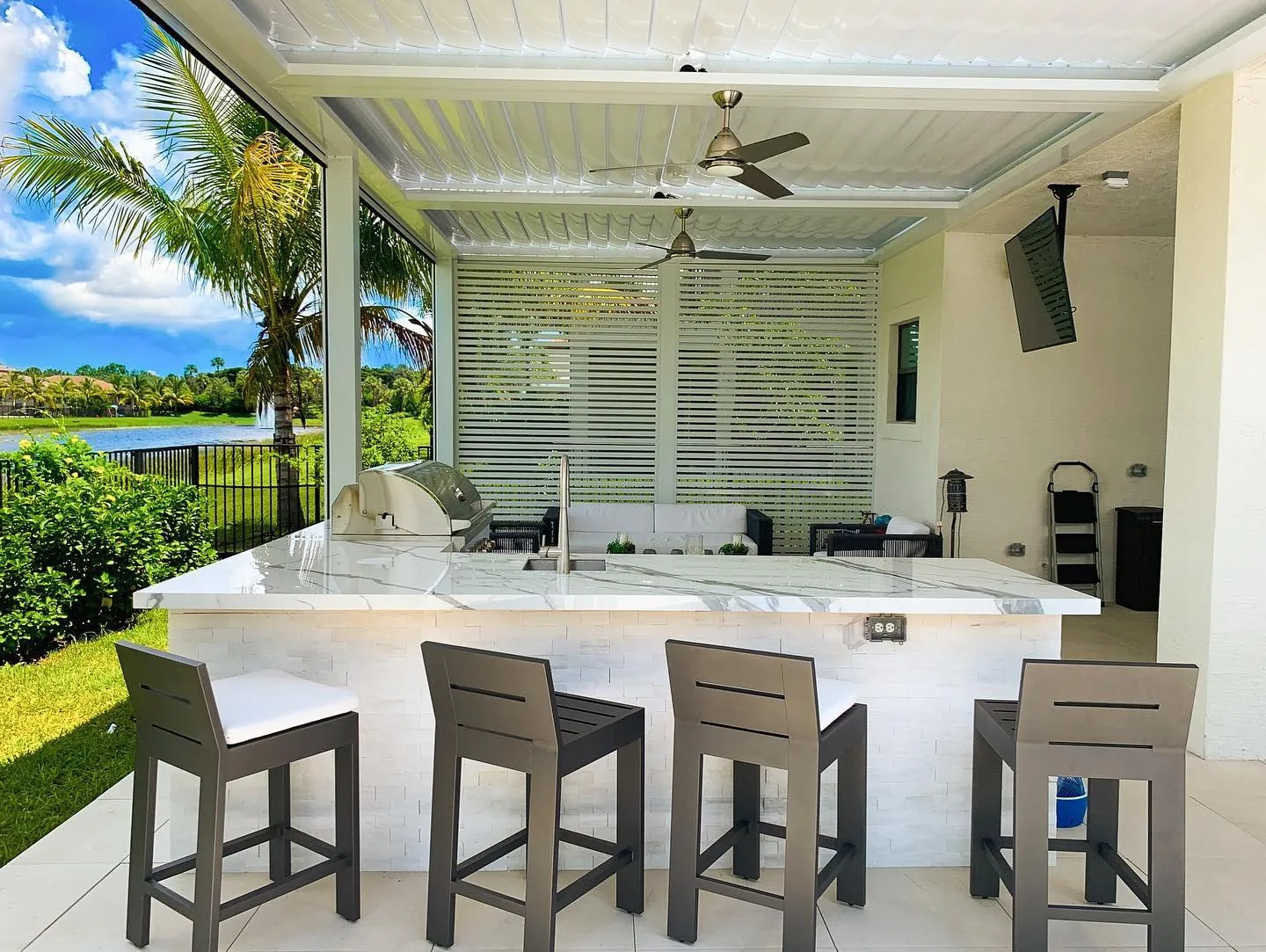 White louvered pergola over a marble-top outdoor kitchen bar with four stools and a lake view.