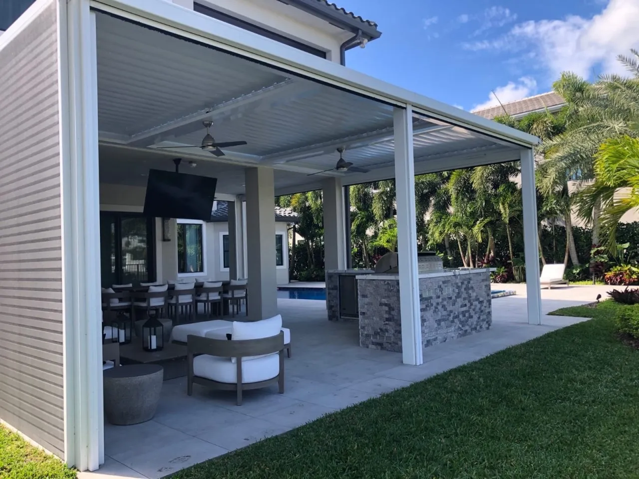 White louvered pergola covering an outdoor kitchen with stacked stone island and lounge seating.