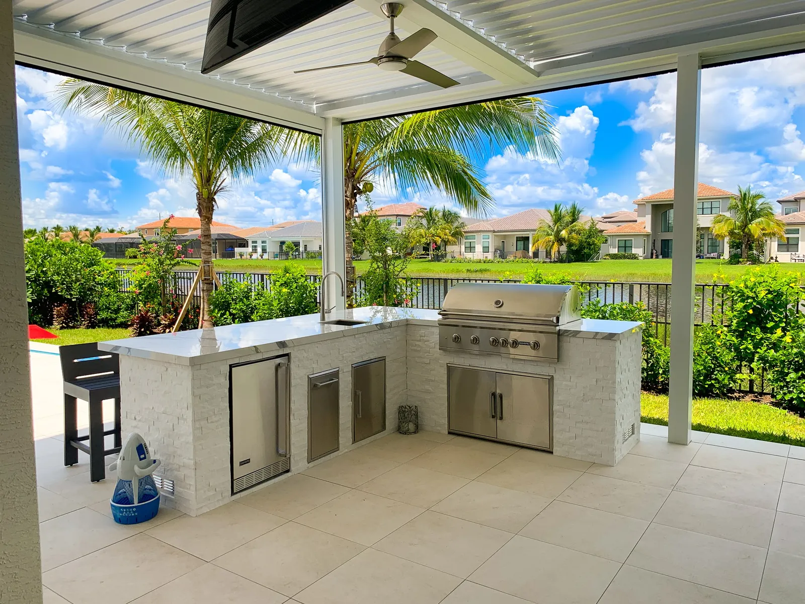 White louvered pergola with ceiling fan over an L-shaped white stone outdoor kitchen by a lake.