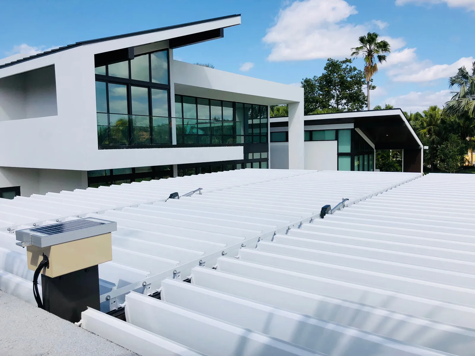 Top of a white louvered roof showing blades, motors and a small solar panel beside a modern home