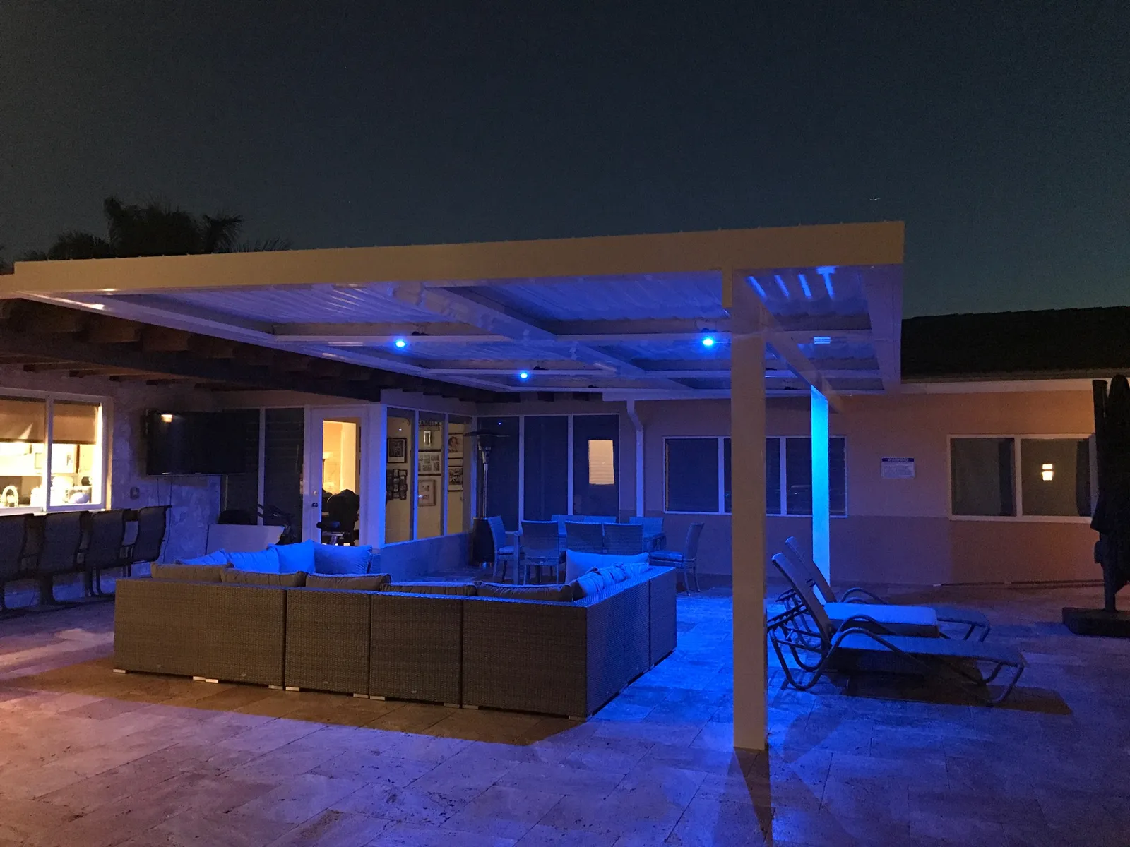 White louvered roof lit with blue LED lights over a travertine patio with wicker seating at dusk
